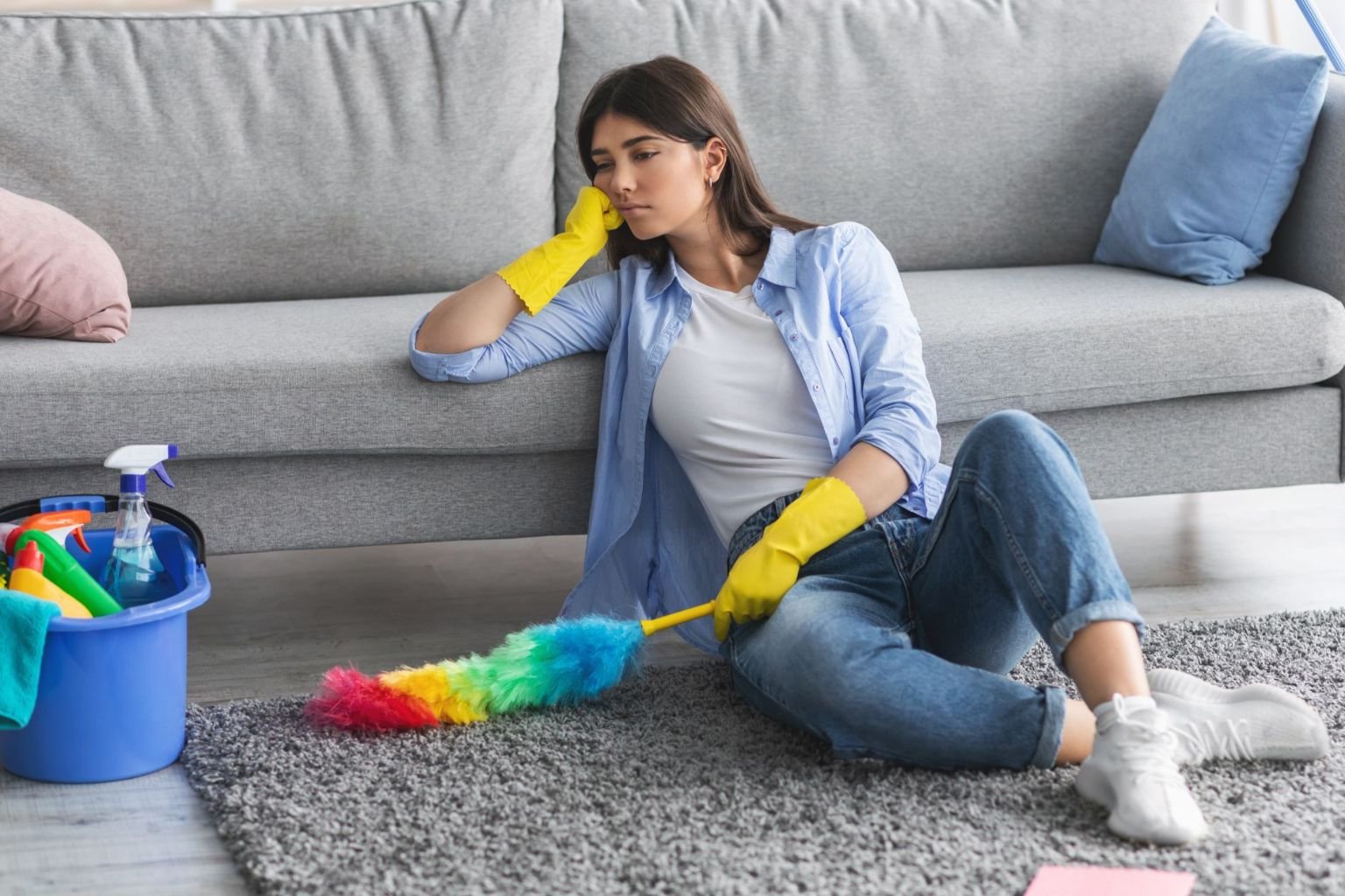 Pensando no que as pessoas sentem antes de descobrir a limpeza cotidiana de carpetes na cansada sentada no chão ao lado de sofá cinza, com luvas amarelas, segurando espanador colorido durante tentativa de limpeza manual de carpete. Balde de produtos de limpeza ao lado mostra o esforço da limpeza caseira tradicional.