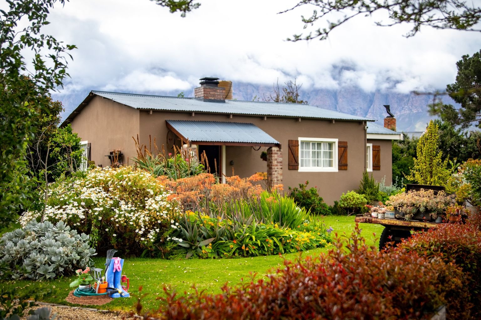Transforme Seu Espaço Verde em um Oásis Sustentável: casa de campo aconchegante com paredes bege, telhado azul de metal e jardim exuberante cheio de flores brancas, laranjas, suculentas e arbustos coloridos. Ferramentas de jardinagem e botas azuis no gramado indicam cuidado ecológico diário, com majestosas montanhas ao fundo sob céu nublado.